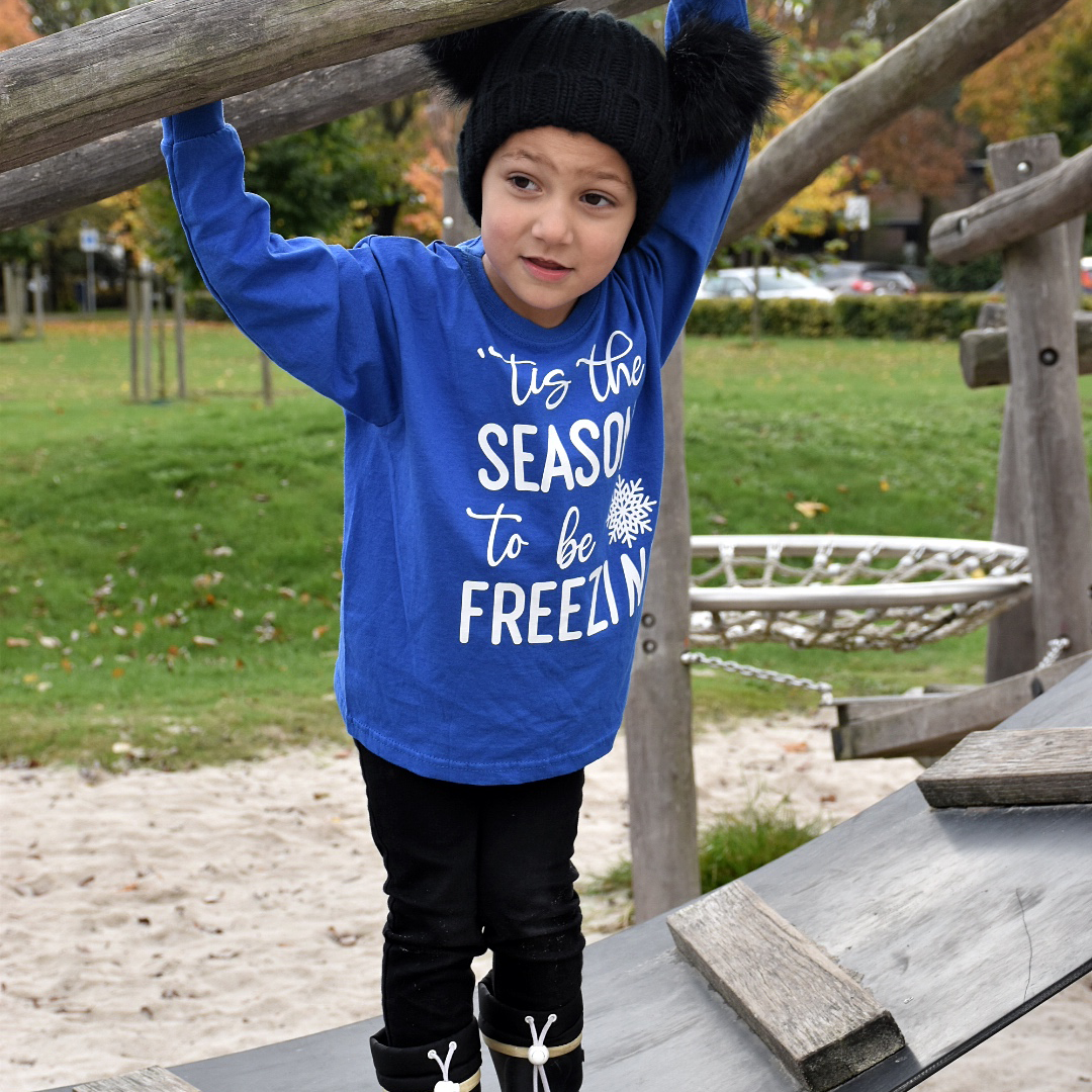 Boy at the playground wearing black hat with pompoms and blue shirt with long sleeves with ''tis the season to be freezin'' print by KMLeon.