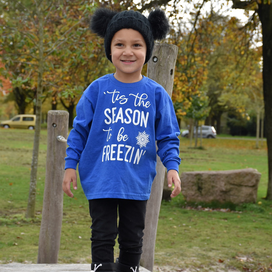 Boy in nature wearing black hat with pompoms and blue shirt with long sleeves with ''tis the season to be freezin'' print by KMLeon.