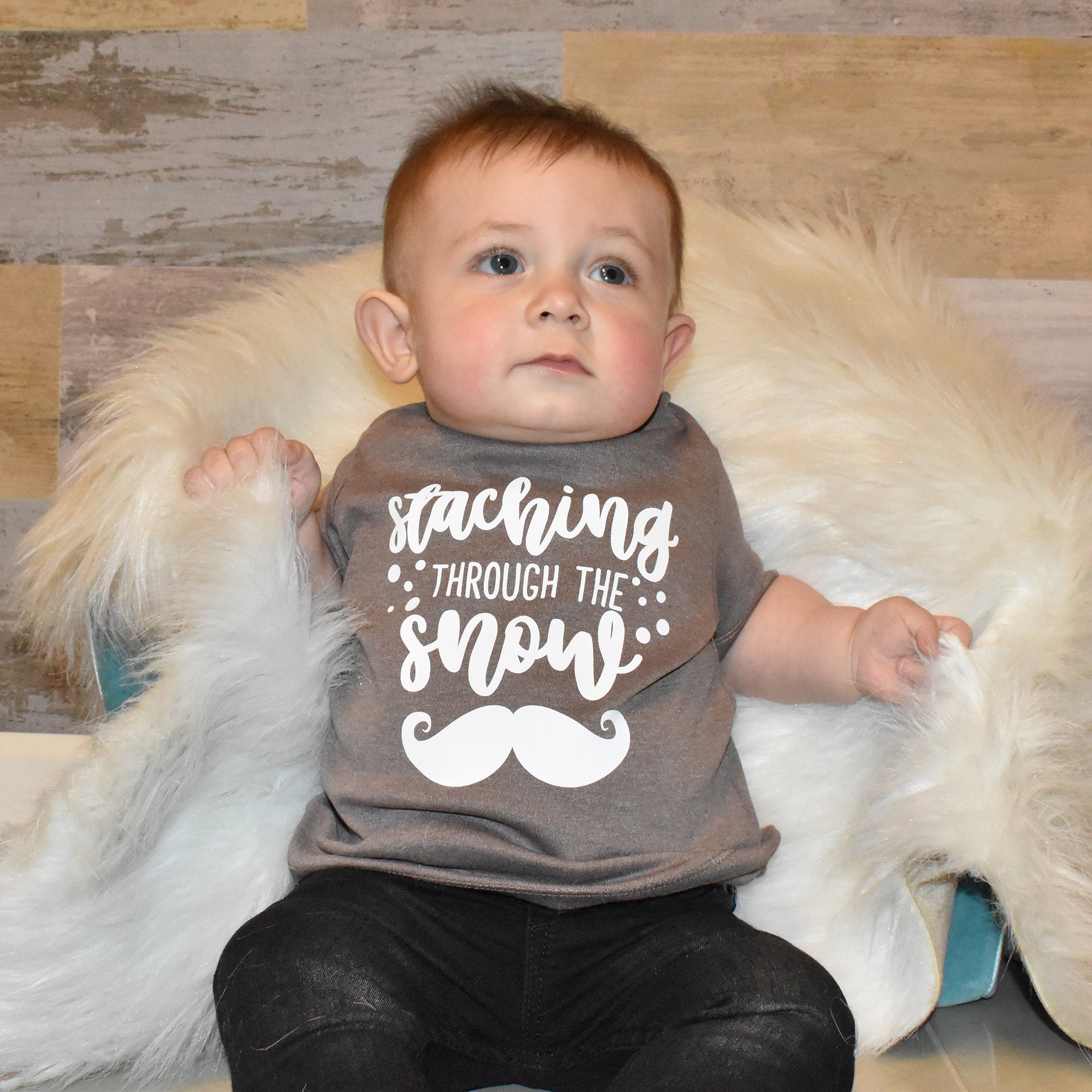 Toddler boy wearing mocha shirt, with 'Staching through the snow' print by KMLeon, leaning against rug.