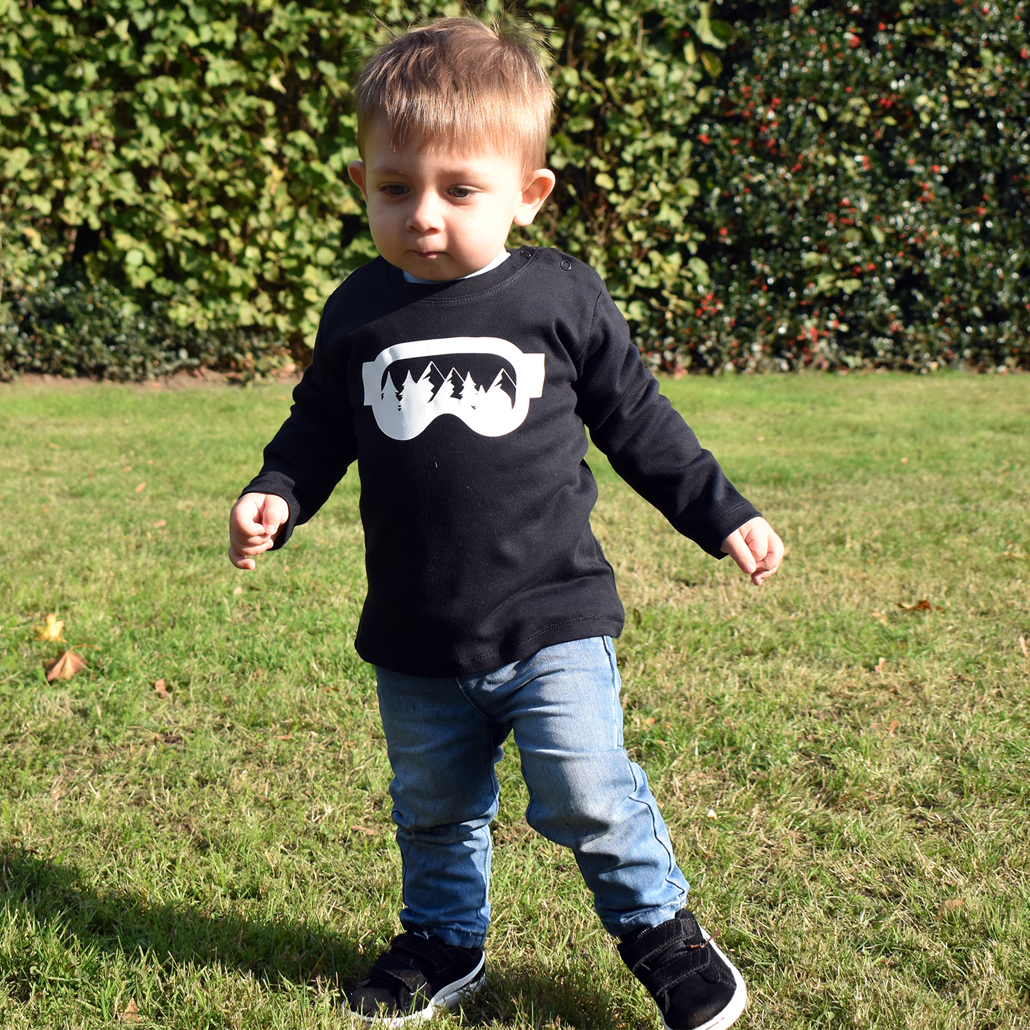 Little boy wearing black shirt with longs sleeves withh ski goggles print by KMLeon walking in park.