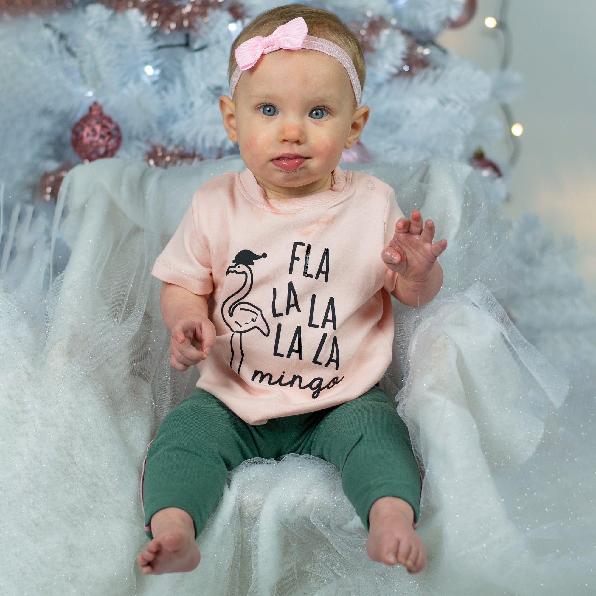 Baby girl with blue eyes, wearing a ppink headband and pink shirt, with 'Fla la la la la mingo' print by KMLeon.