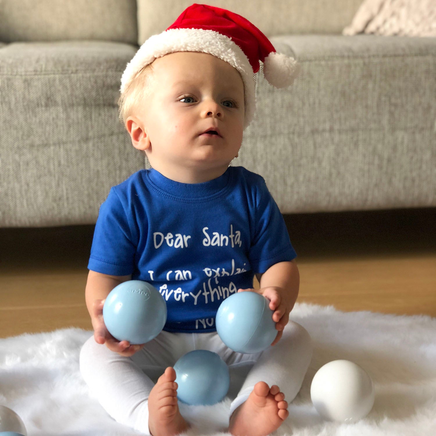 Blonde boy with blue shirt with 'Santa, I can eplain everything' print by KMLeon, playing with christmas ornaments, wearing santa hat.