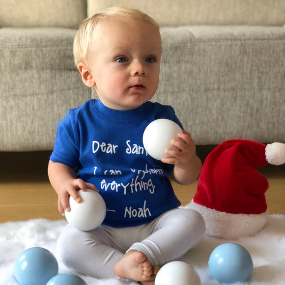 Blonde boy with blue shirt with 'Santa, I can eplain everything' print by KMLeon, playing with christmas ornaments.