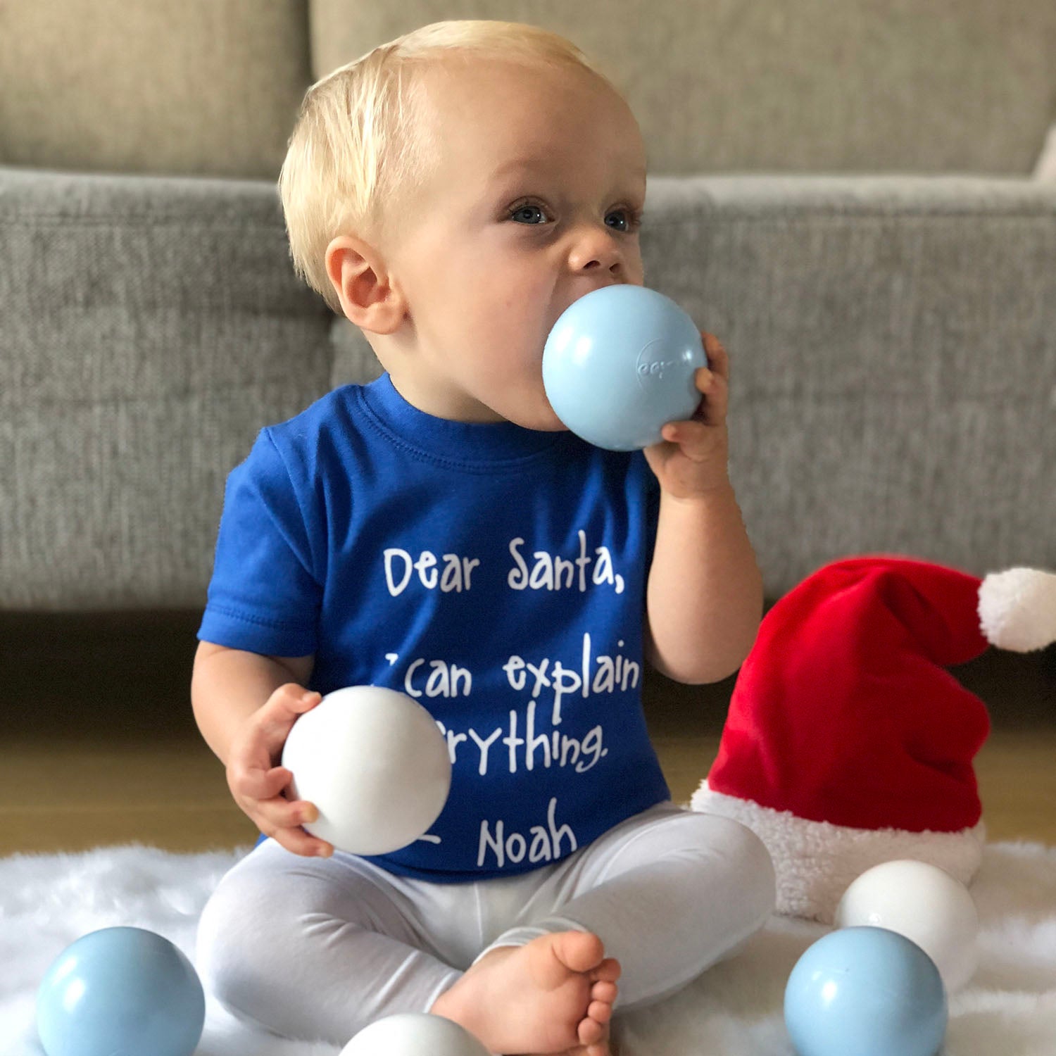 Blonde boy with blue shirt with 'Santa, I can eplain everything' print by KMLeon, putting christmas ornament in mouth.