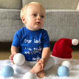 Blonde boy with blue shirt with 'Santa, I can eplain everything' print by KMLeon, playing with christmas ornaments.