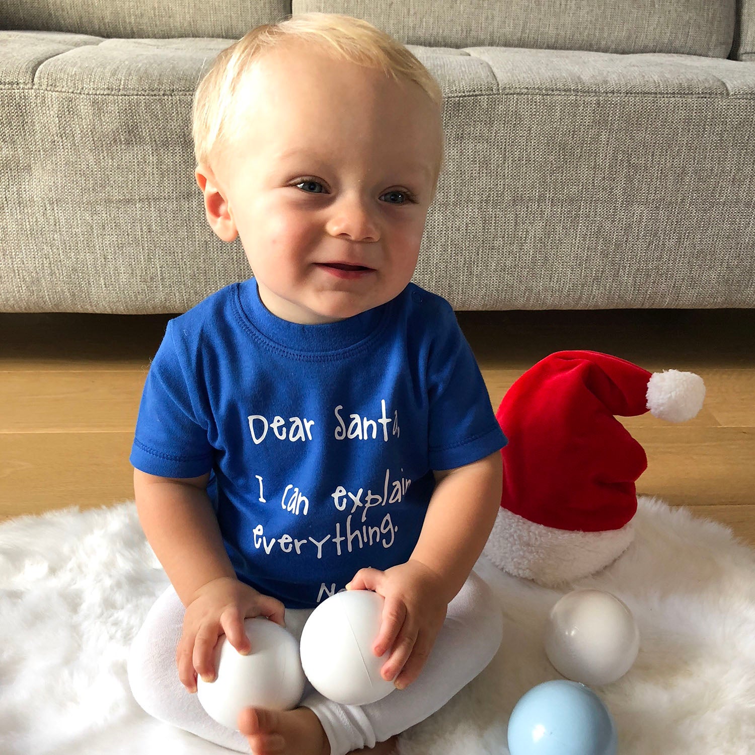 Blonde boy with blue shirt with 'Santa, I can eplain everything' print by KMLeon, playing with christmas ornaments, smiling.