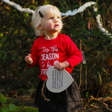 Blonde girl in forest looking up while holding wooden heart wearing black skirt and red shirt with long sleeves with ''tis the season to be freezin'' print by KMLeon.