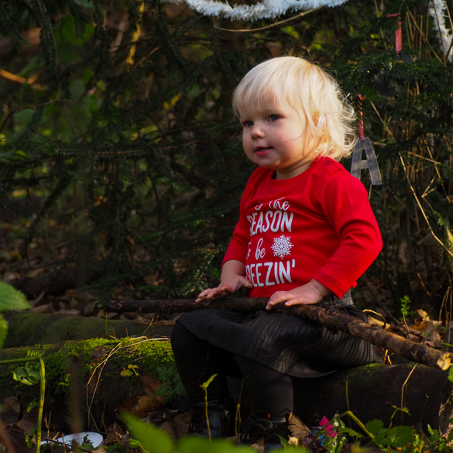 Blonde girl sitting in forest wearing black skirt and red shirt with long sleeves with ''tis the season to be freezin'' print by KMLeon.