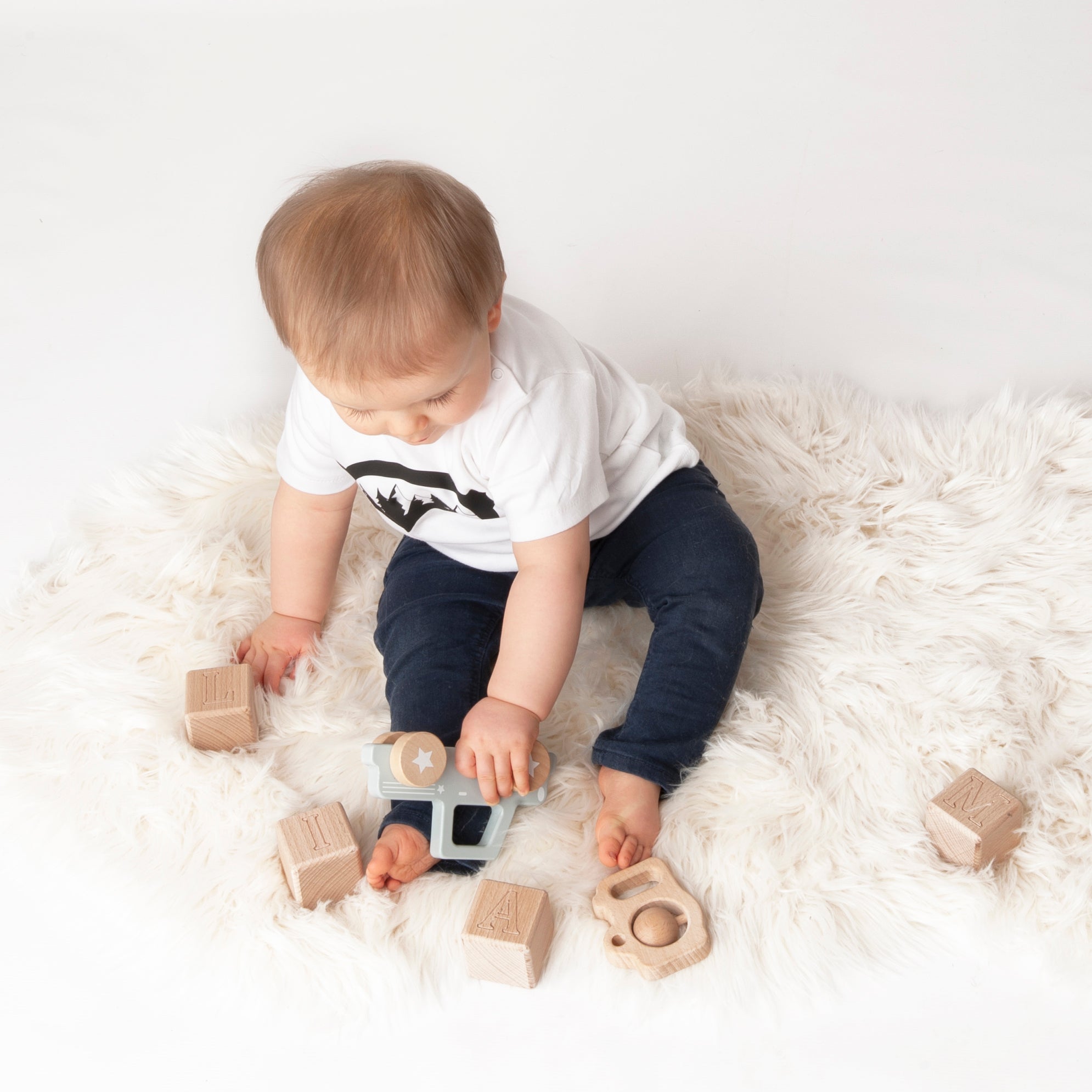 Little boy wearing white shirt with ski goggles print by KMLeon playing with wooden toys.