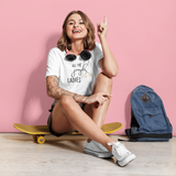 Smiling tattooed woman on yellow skateboard wearing white shirt with 'All the jingle ladies' print by KMLeon, in front of pink wall, pointing her finger up.